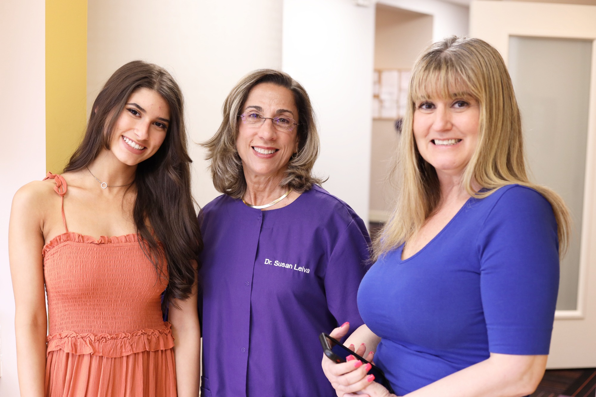 Dr. Susan Leiva of Monroe Orthodontics, in purple scrubs, stands with a smiling patient and a team member in a blue top at the Monroe Township, NJ office.