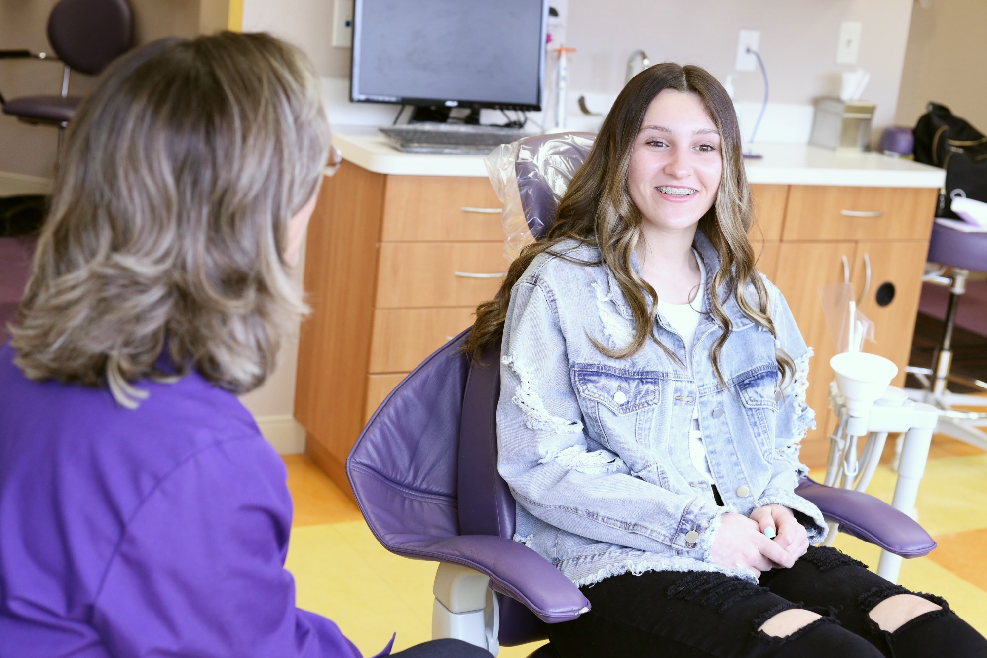 Teen patient in a denim jacket smiling in a purple treatment chair while talking with a team member at Monroe Orthodontics, Dr. Susan Leiva’s Monroe Township orthodontics office