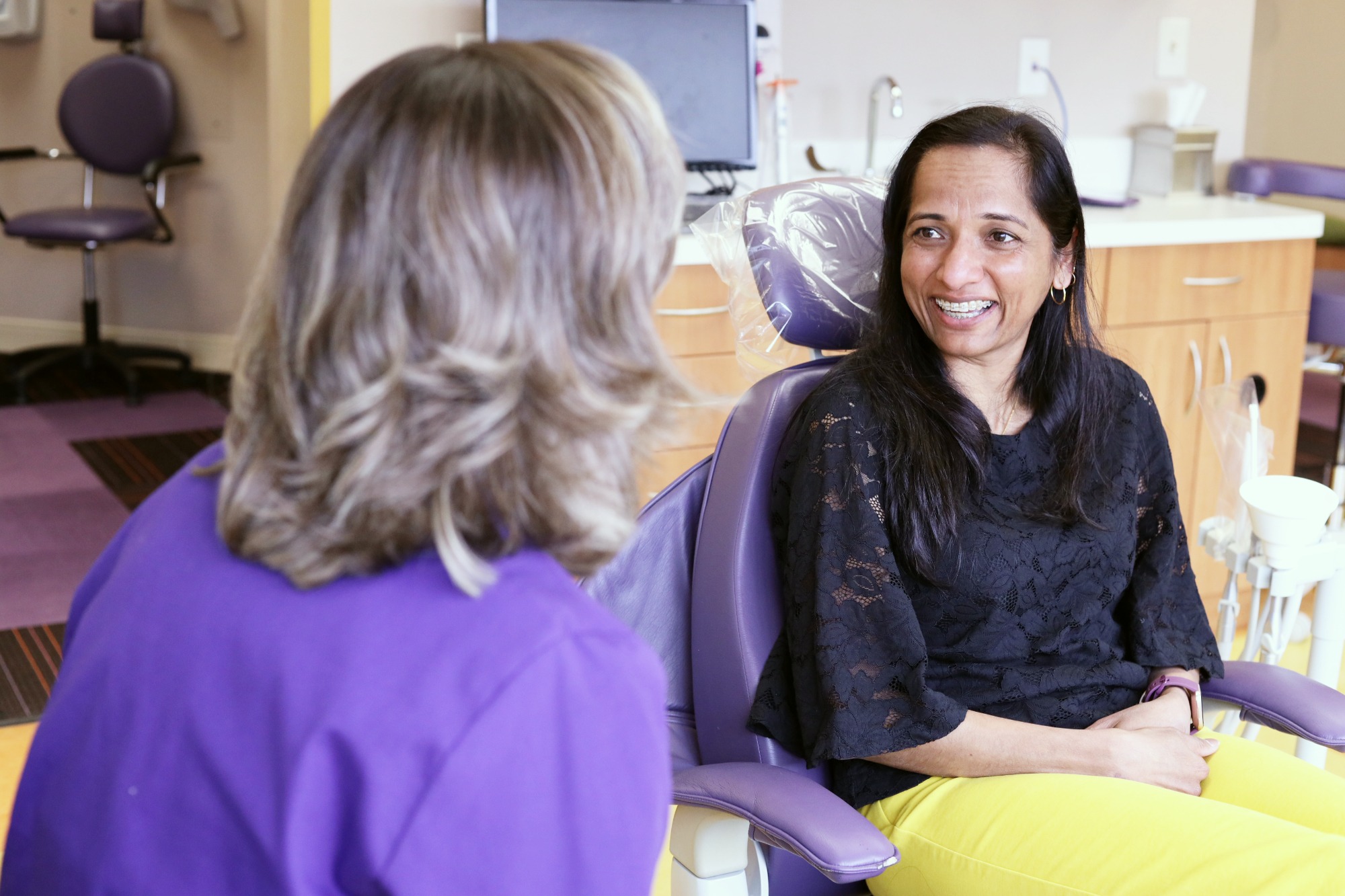 Smiling parent in a black top sits beside the treatment chair and talks with a Monroe Orthodontics team member in purple scrubs during a visit with Dr. Susan Leiva