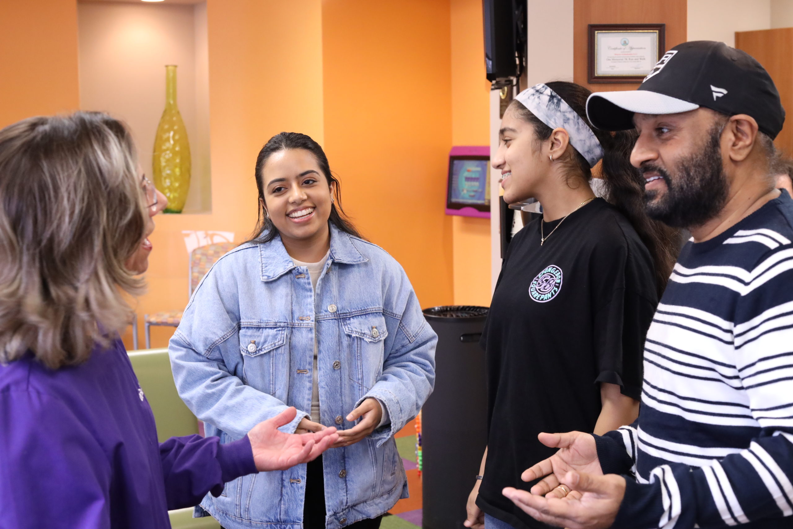 Smiling woman in a denim jacket and a young patient in a black shirt and headband chat in the bright, orange-walled office of Monroe Orthodontics, Dr. Susan Leiva’s practice